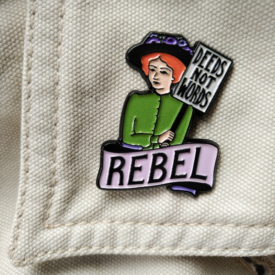 Photo of an enamel pin, depicting a suffragette with ginger hair, a black hat, and a green top. She holds a sign that says “Deeds Not Words” and has a banner beneath her that says “Rebel” in black text on a pink background.