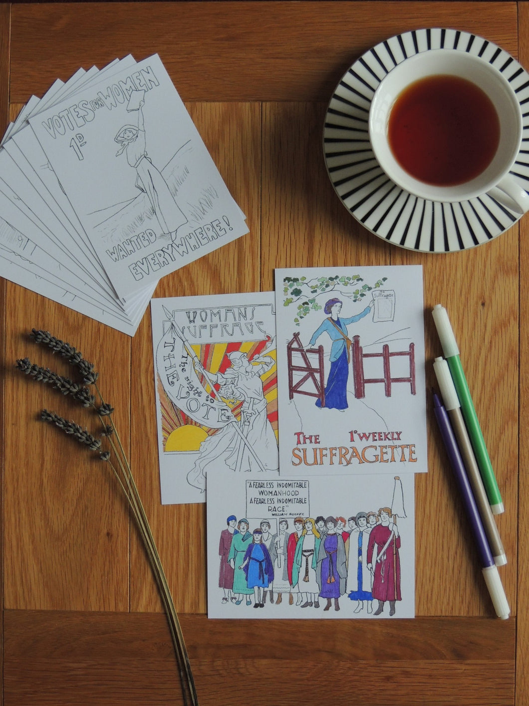Photo of the postcard pack, showing a pile of uncoloured drawings and three coloured. The illustrations are of a suffragette crowd, Lady Liberty wielding a trumpet and sword and banner, and a suffragette holding a newsletter on behalf of the cause. Next to the cards are three felt tip pens, a sprig of lavender, and a cup and saucer of black tea.