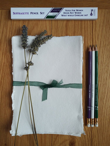 Set of three graphite pencils pictured next to handmade paper and a sprig of lavender. The pencils are painted purple, green & white, featuring slogans 