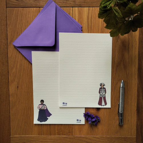 Photo of two pieces of notepaper alongside a purple envelope, a pen, and some leaves. The pages are lined and each have an illustrated suffragette in colour in the corners, one of whom holds a sign saying “Never Give Up The Fight.”