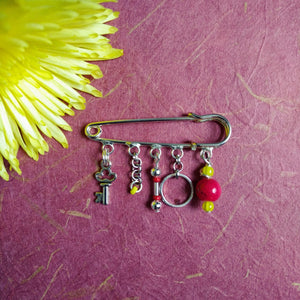 Photo of the silver brooch from which dangles a small key charm, interlinked chains, a ring, and red and yellow beads. Next to the brooch are the petals of a yellow flower.