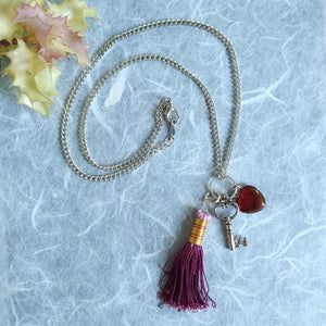Photo of a necklace with a silver chain that ends in a violet tassel, Scarlett glass bead, and a small silver key charm. Next to the necklace is some green leaves.