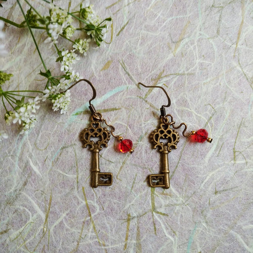 Photo of two key-shaped earrings with ornate designs, both linked with a scarlet glass bead. The earrings are next to small white flowers with green stems.