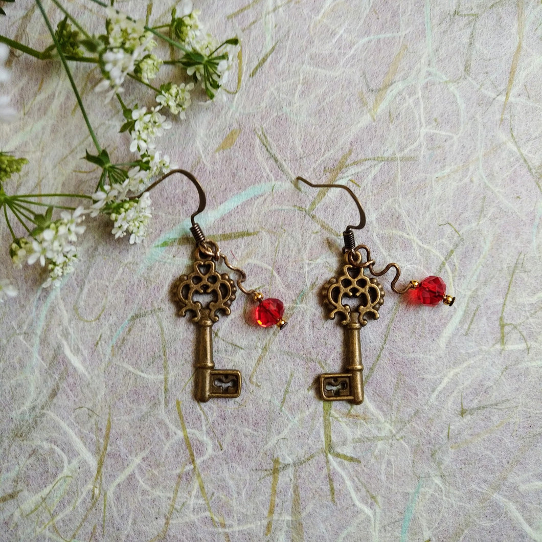 Photo of two key-shaped earrings with ornate designs, both linked with a scarlet glass bead. The earrings are next to small white flowers with green stems.