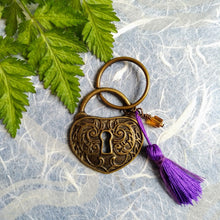 Load image into Gallery viewer, Photo of a bronze-coloured keyring that features a heart-shaped lock with a keyhole at its centre, joined with a violet tassel and amber-coloured bead. Next to the keyring are some green leaves.