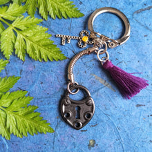Load image into Gallery viewer, Photo of black heart-shaped lock with keyhole keyring, along with a smaller key charm, amber-coloured bead, and a violet tassel. Next to the keyring are some green leaves.
