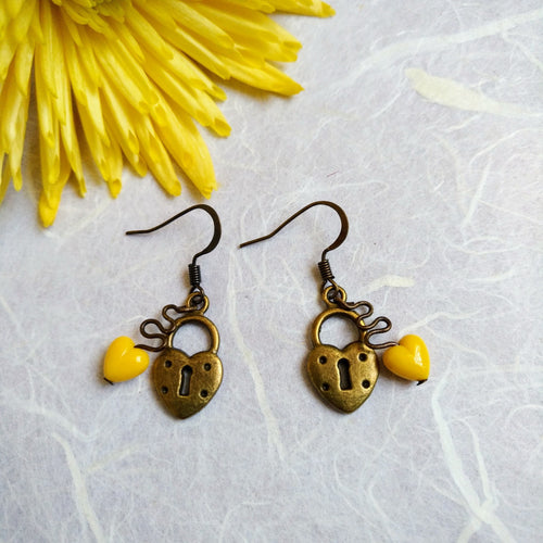 Photograph of two earrings which depict locks in the shape of hearts, with keyholes in the centre. Attached to each one is a smaller yellow heart, by a winding link. Next to the earrings are some yellow petals.