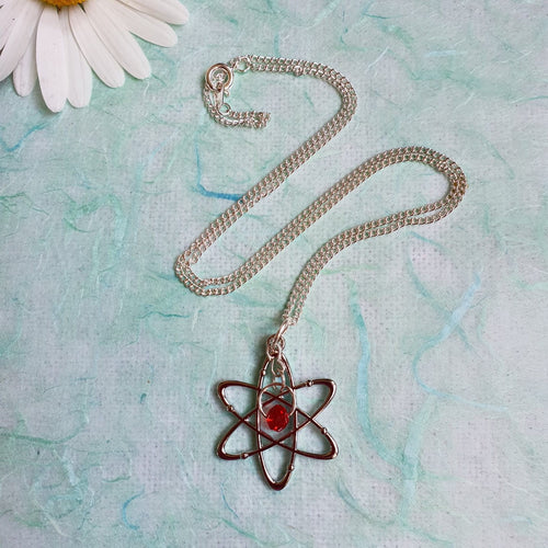 Photograph of a necklace with an atomic structure pendant with a scarlet glass bead at the centre, attached to a silver chain. Pictured with a white flower.