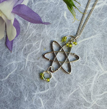 Load image into Gallery viewer, Photo of a silver necklace. The chain is joined to a pendant in the shape of an atomic structure with green “electron” rings at the top and bottom. Next to the necklace are some lilac and white petals.