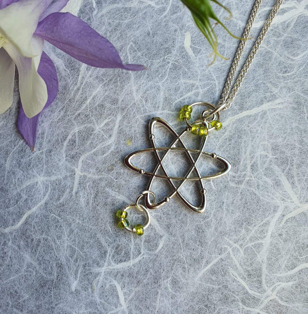 Photo of a silver necklace. The chain is joined to a pendant in the shape of an atomic structure with green “electron” rings at the top and bottom. Next to the necklace are some lilac and white petals.
