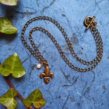 Load image into Gallery viewer, A chain necklace laid out in a wave shape, with a fleur-de-lis pendant and a little pearl bead suspended from the bottom. The background of blue is a contrast to the antique gold colour of the necklace, and ivy decorates the side of the frame.