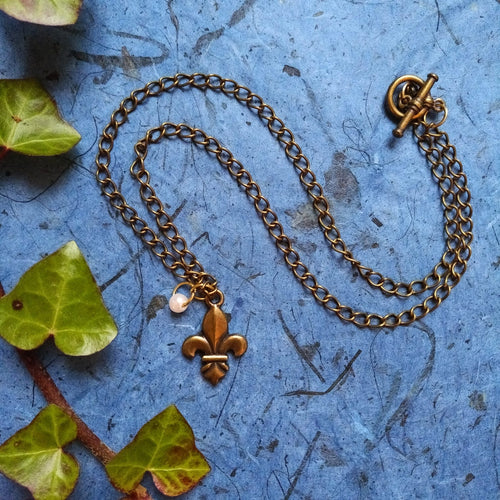A chain necklace laid out in a wave shape, with a fleur-de-lis pendant and a little pearl bead suspended from the bottom. The background of blue is a contrast to the antique gold colour of the necklace, and ivy decorates the side of the frame.