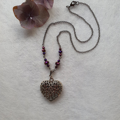 Photograph of a necklace with a heart pendent with intricate metal filligree patterning, and beads either side in different shades of violet and purple. Pictured with a purple flower.