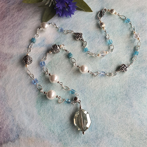 Photo of a necklace next to a purple flower. The necklace chain links are threaded with faux pearls, blue beads, crystals, and end in an oval-shaped embellished metal pendant.