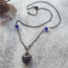 Load image into Gallery viewer, Photo of a necklace with a silver chain interlinked with royal blue glass beads. The locket is shaped like a heart with embellishments on the surface. Next to the necklace are some faintly dark-red leaves.