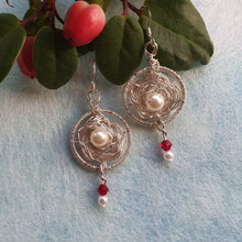 Load image into Gallery viewer, Photo of two earrings, circles of crocheted wire roses with faux pearls at the centre. Scarlet glass crystals hang from the base. Next to the earrings are some leaves and red berries.