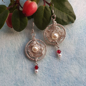 Photo of two earrings, circles of crocheted wire roses with faux pearls at the centre. Scarlet glass crystals hang from the base. Next to the earrings are some leaves and red berries.