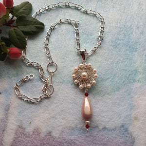 Photo of a necklace with a silver chain. The necklace pendants is a circle of faux pearls with a large one at the centre, and a tear-shaped piece of acrylic that falls from the base. Next to the necklace are some leaves and red buds.