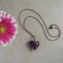 Load image into Gallery viewer, Photo of a necklace with a silver chain that ends in a purple glass heart with a silver adornment. Next to the necklace is a pink flower.