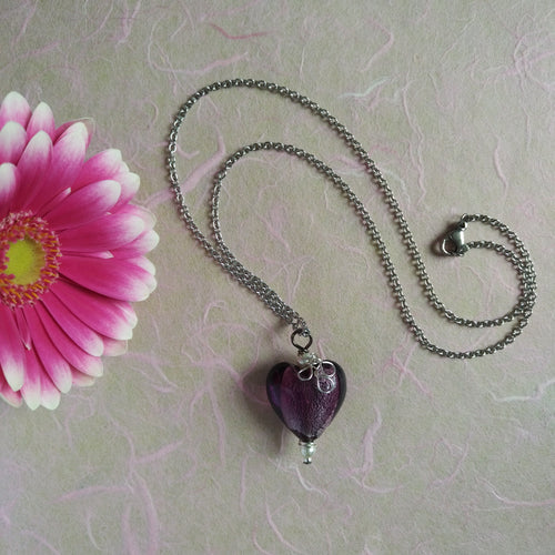 Photo of a necklace with a silver chain that ends in a purple glass heart with a silver adornment. Next to the necklace is a pink flower.