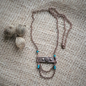 Photo of a necklace with a copper tone chain. The chain threads through a metal bar with three small circles in the centre, the middle of which contains a spiral design. There are small turquoise metal adornments on the chain. Next to the necklace are some seed pods.