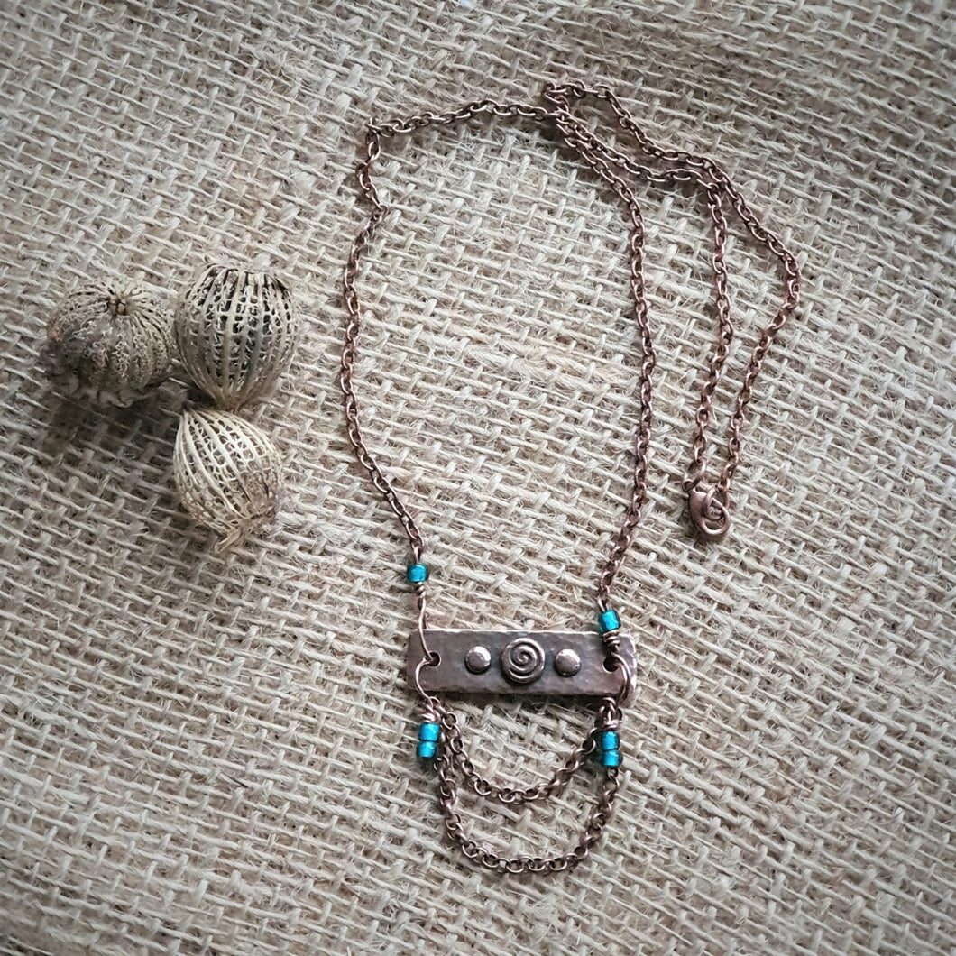 Photo of a necklace with a copper tone chain. The chain threads through a metal bar with three small circles in the centre, the middle of which contains a spiral design. There are small turquoise metal adornments on the chain. Next to the necklace are some seed pods.