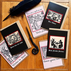 Photo of a the “Witches’ Coven” card pack with all six laid out next to each other in a different configuration, showing the “Best Fiends Forever” card alongside “Witches Be Blazin’”, “Party like its 1599”, “Haters Gonna Hate”, “Hex Addict”, and “Witch, Please!”. Along with the brown envelope, also pictured are a black quill and a letter opening with a curled end.