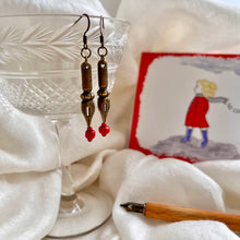 Load image into Gallery viewer, Photo of the the earrings dangling from a crystal glass. They are both bronze and shaped like pen nibs. Red beads are fixed at the tip. Resting nearby are a fountain pen and an illustrated drawing of a girl on the moors with her scarf blowing in the wind.