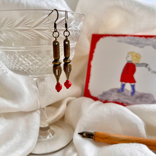 Photo of the the earrings dangling from a crystal glass. They are both bronze and shaped like pen nibs. Red beads are fixed at the tip. Resting nearby are a fountain pen and an illustrated drawing of a girl on the moors with her scarf blowing in the wind.