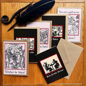 Photo of a the “Witches’ Coven” card pack with all six laid out next to each other, showing the “Best Fiends Forever” card alongside “Witches Be Blazin’”, “Party like its 1599”, “Haters Gonna Hate”, “Hex Addict”, and “Witch, Please!”. Along with the brown envelope, also pictured are a black quill and a letter opening with a curled end.