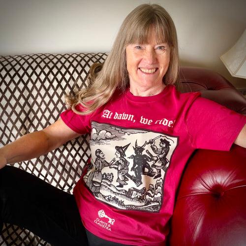 Photograph of a t-shirt modelled by a smiling white-skinned woman with long silvery brown hair. The cherry red coloured t-shirt features a monochrome mediaeval woodcut design depicting a woman addressing witches and a demon each mounted on broomsticks. Above in Old English text are the words 