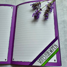 Load image into Gallery viewer, Photo from inside the notebook, with blank lined pages framed by purple colouring. In the bottom right corner is a sash that reads “Votes For Women”. On the notebook are some sprigs of lavender.