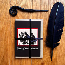 Load image into Gallery viewer, Photo of a the “Witches’ Coven” card pack wrapped in black ribbon, showing the “Best Fiends Forever” card on top. Along with the brown envelope, also pictured are a black quill and a letter opening with a curled end.
