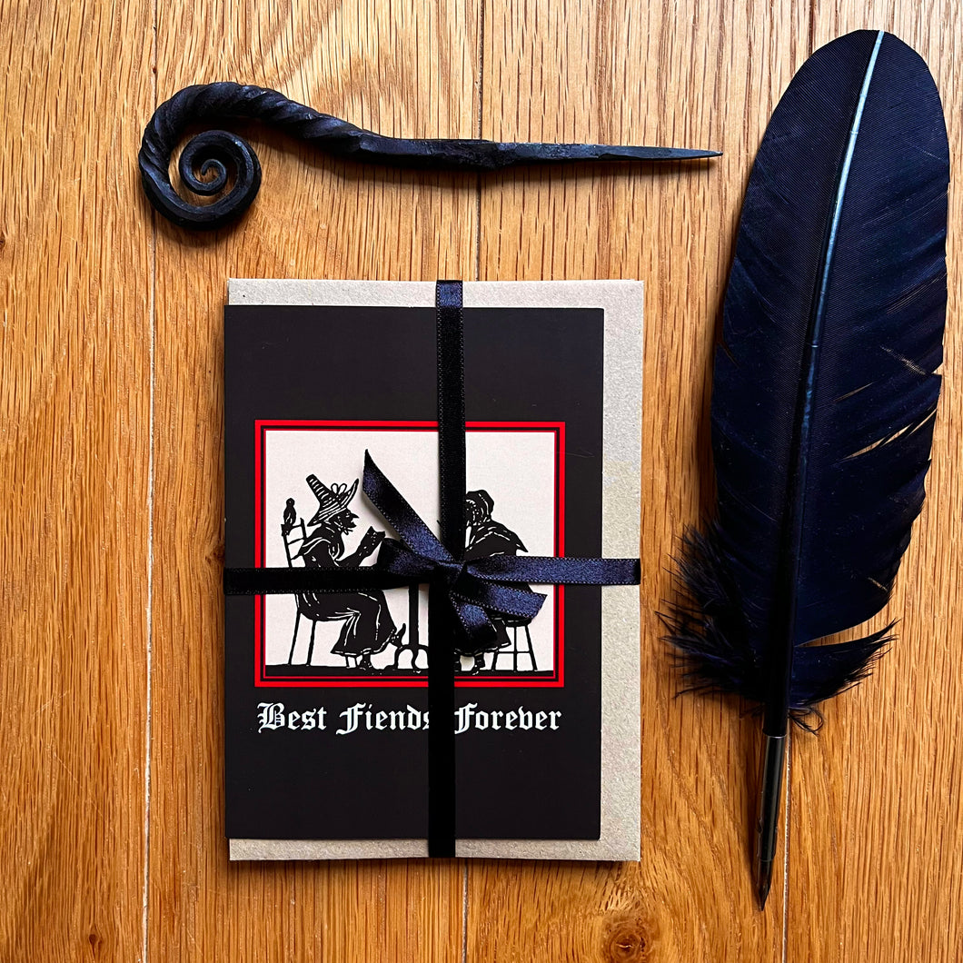Photo of a the “Witches’ Coven” card pack wrapped in black ribbon, showing the “Best Fiends Forever” card on top. Along with the brown envelope, also pictured are a black quill and a letter opening with a curled end.