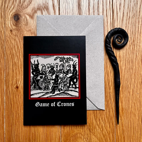 Photo of a black coloured card featuring a mediaeval woodcut artwork depicting witches and demons drinking and celebrating around a table. Beneath it the caption reads 'Game of Crones'. Pictured with a brown envelope and black letter opener with a curved, spiral end.