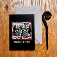 Photo of a black coloured card featuring a mediaeval woodcut artwork depicting witches and demons drinking and celebrating around a table. Beneath it the caption reads 'Game of Crones'. Pictured with a brown envelope and black letter opener with a curved, spiral end.