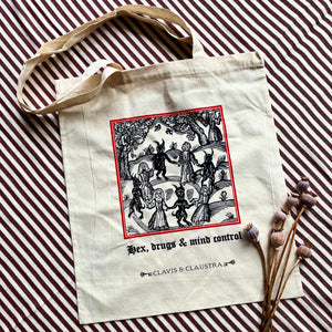 Large cotton tote bag, pictured with a sprig of dried poppies on a striped background. The bag features a Mediaeval woodcut illustration in black & scarlet of people dancing in a circle with devils, with "Hex, drugs and mind control" written underneath in Gothic typeface.