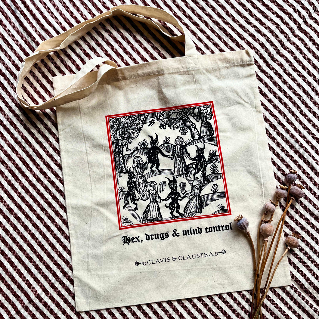 Large cotton tote bag, pictured with a sprig of dried poppies on a striped background. The bag features a Mediaeval woodcut illustration in black & scarlet of people dancing in a circle with devils, with 
