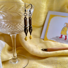 Load image into Gallery viewer, Photo of the the earrings dangling from a crystal glass. They are both bronze and shaped like pen nibs. Yellow beads are fixed at the tip. Resting nearby are a fountain pen and an illustrated drawing of a girl on the moors with her scarf blowing in the wind.