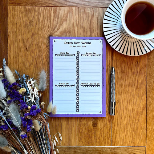 A photo of a notepad with a pen, a bunch of dried flowers and a cup of black tea. The pad has a purple border and is entitled 'Deeds Not Words' with the page sectioned off into 'Must do', 'Should do', 'Could do' and 'Would like to do', decorated with chains and bunting.