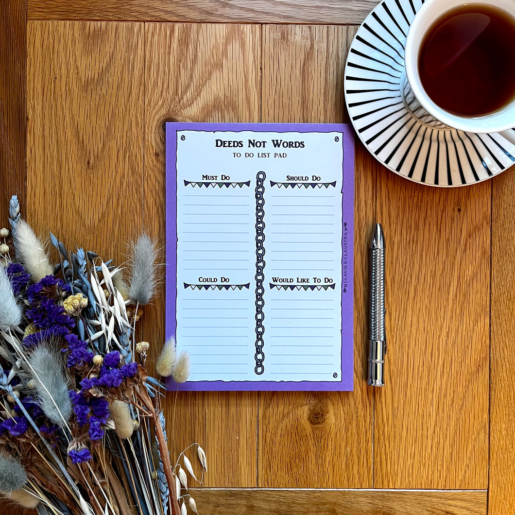 A photo of a notepad with a pen, a bunch of dried flowers and a cup of black tea. The pad has a purple border and is entitled 'Deeds Not Words' with the page sectioned off into 'Must do', 'Should do', 'Could do' and 'Would like to do', decorated with chains and bunting.