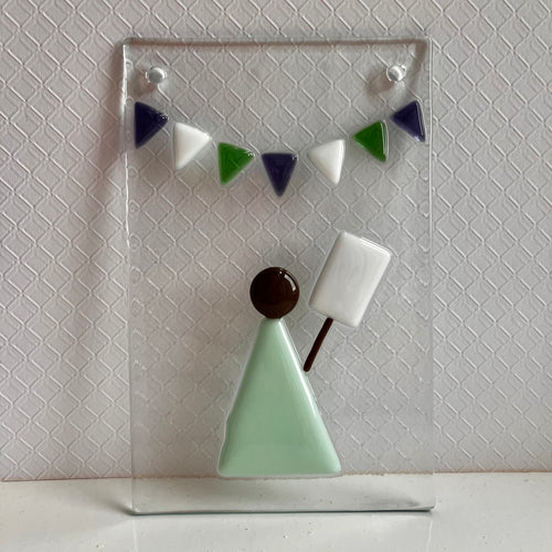 Photo of glass artwork, a rectangle that shows a suffragette in a minimalist style holding a sign beneath some white, green, and purple bunting. Two holes for hanging the artwork are in the top two corners.