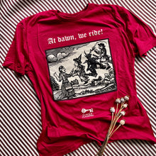 Load image into Gallery viewer, Photograph of a t-shirt laid out on a burgundy &amp; white striped fabric, next to some dried poppy heads. The cherry red coloured t-shirt features a monochrome mediaeval woodcut design depicting a woman addressing witches and a demon each mounted on broomsticks. Above in Old English text are the words &quot;At dawn we ride&quot;.