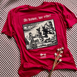 Photograph of a t-shirt laid out on a burgundy & white striped fabric, next to some dried poppy heads. The cherry red coloured t-shirt features a monochrome mediaeval woodcut design depicting a woman addressing witches and a demon each mounted on broomsticks. Above in Old English text are the words "At dawn we ride".