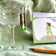 Photo of the the earrings dangling from a crystal glass. They are both bronze and shaped like pen nibs. Green beads are fixed at the tip. Resting nearby are a fountain pen and an illustrated drawing of a girl on the moors with her scarf blowing in the wind.