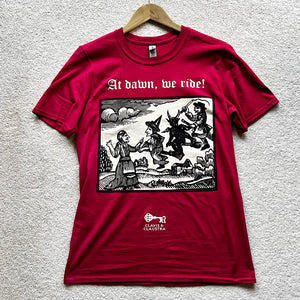 Photograph of a t-shirt hung on a coathanger and laid out on a pale background. The cherry red coloured t-shirt features a monochrome mediaeval woodcut design depicting a woman addressing witches and a demon each mounted on broomsticks. Above in Old English text are the words "At dawn we ride".