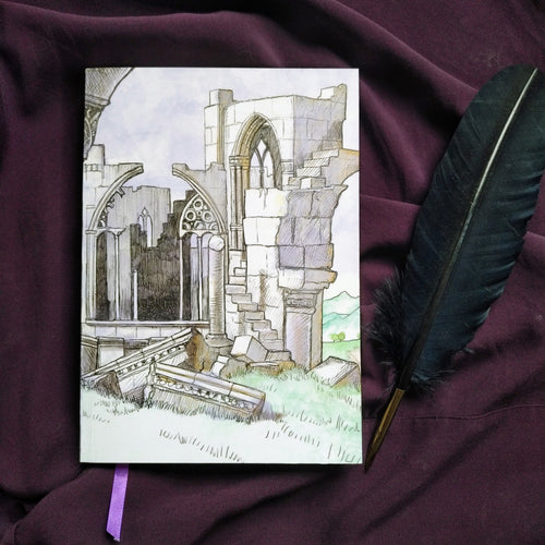 Photo of the Thornfield Hall notebook, depicting a crumbling Gothic ruin with a purple ribbon emerging from the pages, with walls and columns  and windows broken into rocks on the grass. Next to the book is a black quill.