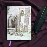 Photo of the Thornfield Hall notebook, depicting a crumbling Gothic ruin with a purple ribbon emerging from the pages, with walls and columns  and windows broken into rocks on the grass. Next to the book is a black quill.