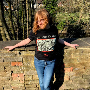 Photo of a white woman outside and smiling, leaning against a wall. She wears blue jeans and a black t-shirt featuring a black-and-white mediaeval woodcut artwork on a black background and framed in red, depicting several demons cavorting around a gigantic ring. One plays some bagpipes, another the lute. Above it the caption reads 'Party like it’s 1599' in a white Gothic typeface. Underneath is the Clavis & Claustra logo in red.